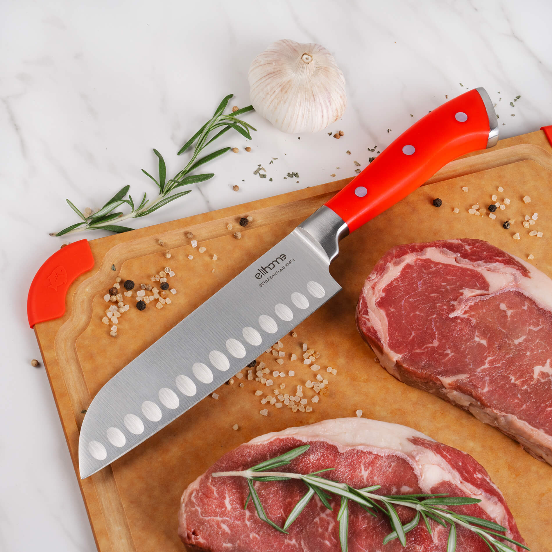 Elihome santoku knife with red handle on wood fiber cutting board, displayed with raw ribeye steaks, garlic, rosemary, and pink salt