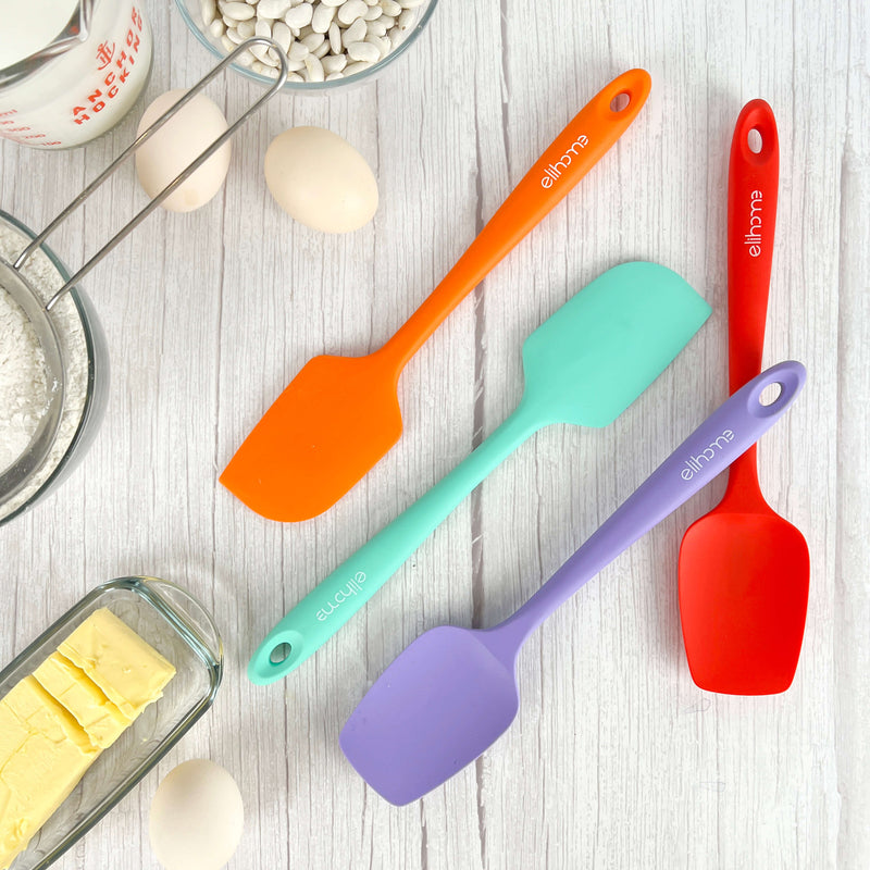 Elihome silicone spatulas in orange, mint, purple, and red displayed with baking ingredients including eggs, butter, flour, and measuring cup on white wood background