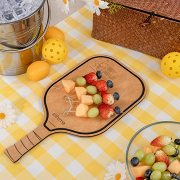 Fruit salad on a paddle-shaped cutting board with a yellow checkered tablecloth, picnic basket, and lemon on a wooden surface.