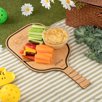 Vegetable platter with hummus on a wooden paddle, set on a picnic blanket with flowers and a basket.