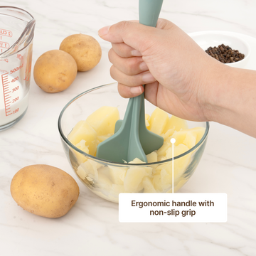 Hand mashing boiled potatoes in a glass bowl with an Elihome Classic Chopper in Sage Green, ergonomic handle with non-slip grip