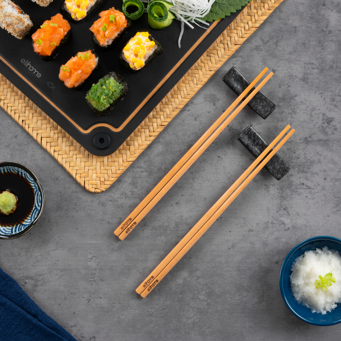 Overhead view of a sushi platter on an Elihome board with two pairs of Elihome wood fiber chopsticks, a dish of wasabi and a bowl of rice on a grey stone surface