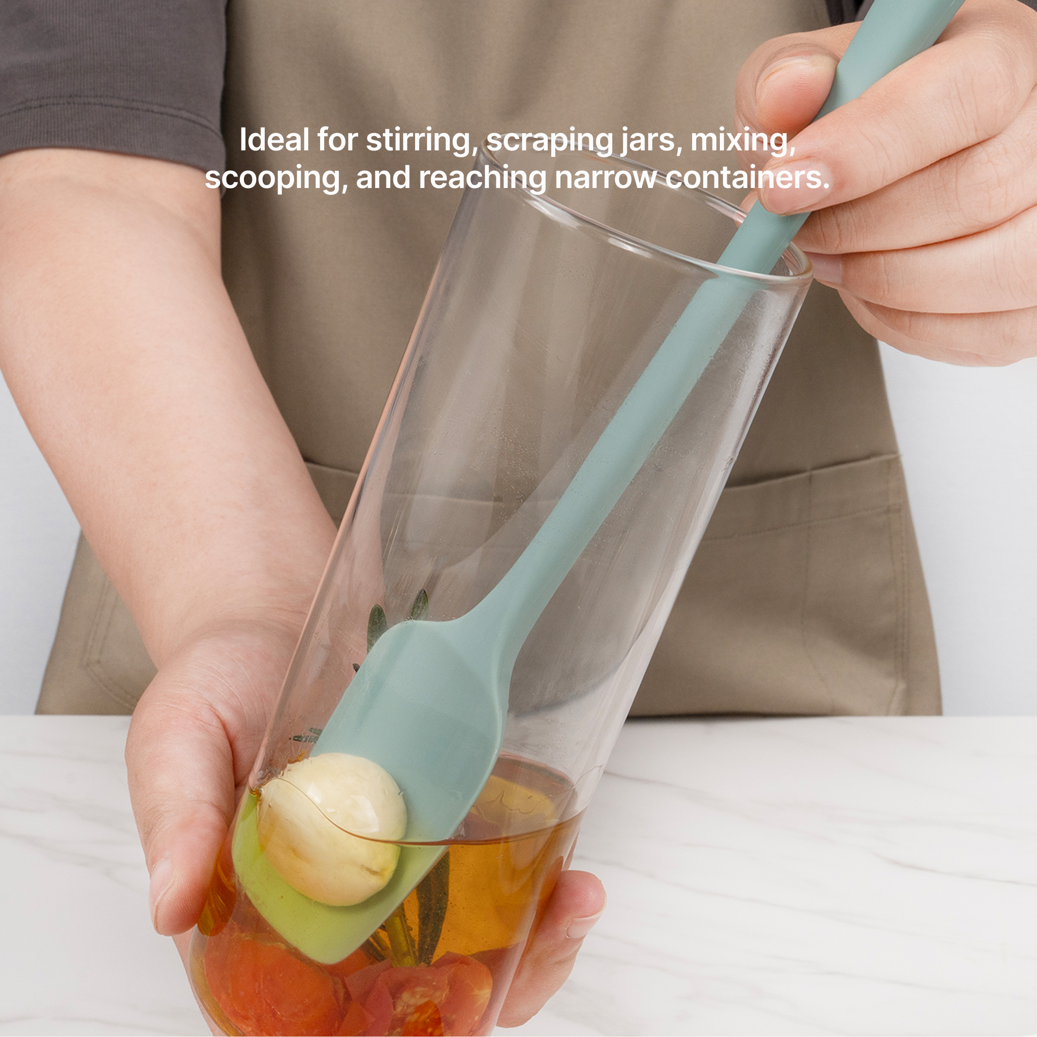 Hand using an Elihome Skinny Slim silicone spoonula in Teal Grey to reach fruit at the bottom of a tall glass, ideal for stirring, scraping jars, mixing, scooping and reaching narrow containers