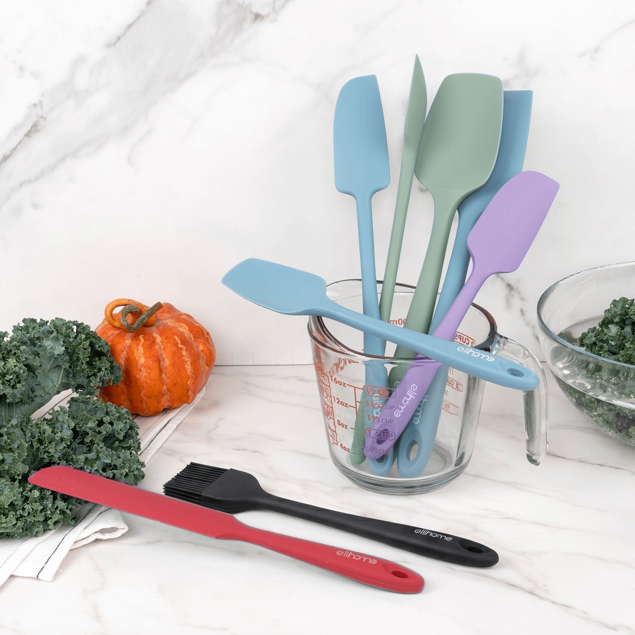 Elihome silicone spatulas and spoonulas in Teal Grey, Sage Green, Purple, Tiffany Blue, Red and Black in a glass measuring cup on a marble counter with kale and a pumpkin
