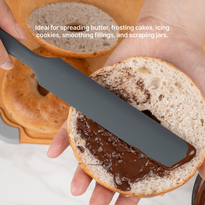 Hand using the Elihome Premium Silicone Spreader in Gray to spread chocolate on a bagel half, ideal for spreading butter, frosting cakes, icing cookies, smoothing fillings and scraping jars